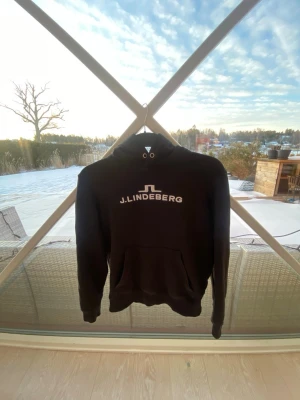 Svart hoodie från J.Lindeberg - Snygg svart hoodie från J.Lindeberg med vit logga och text på bröstet. Klassisk modell med huva och dragsnören samt en stor magficka framtill. Perfekt för en avslappnad och trendig stil. Tillverkad i mjukt material för extra komfort.