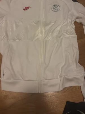 Vit Nike PSG ziptröja - Passar dig som är runt 180 och 75 kg, tröjan är som en small L skulle jag säga. Säljer en vit ziptröja från Nike med PSG-logga på bröstet och röd Nike Swoosh. Tröjan har hel dragkedja, ribbade muddar och är tillverkad i ett mjukt, sportigt material. Perfekt för dig som gillar fotboll och vill ha en clean look.