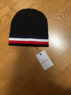 Svart Moncler mössa med logga - Snygg svart mössa från Moncler med klassisk Moncler-logga framtill. Mössan har en uppvikt kant med ränder i blått, vitt och rött som ger en sportig vibe. Perfekt accessoar för kalla dagar och passar till de flesta outfits.