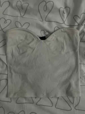 Vit topp från Gina Tricot - Supersnygg vit bandeau topp från Gina Tricot i ribbat material. Toppen är axelbandslös med en hjärtformad byst och croppad passform. Perfekt att styla med höga jeans eller kjol för en trendig look. Använd men i bra skick.