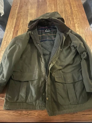 Grön Barbour vindjacka med huva - Säljer en klassisk grön jacka från Barbour med huva och rutigt innerfoder. Jackan har två stora fickor framtill, dragkedja och tryckknappar. Materialet är vaxad bomull som ger en tålig och vattenavvisande yta. Perfekt för dig som gillar tidlös brittisk stil. Nypris 6900:- 