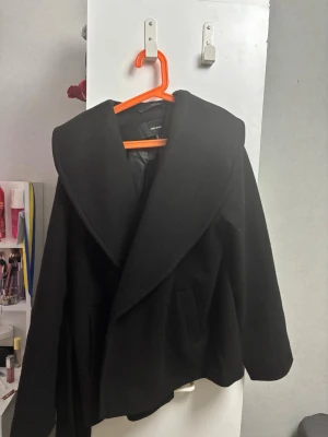 Svart kappa från Vero Moda - Stilren svart kappa från Vero Moda med bred, markerad krage och knytskärp i midjan. Kappan har en rak passform och är tillverkad i ett mjukt, ull-liknande material. Perfekt för dig som vill ha en klassisk look med modern twist.