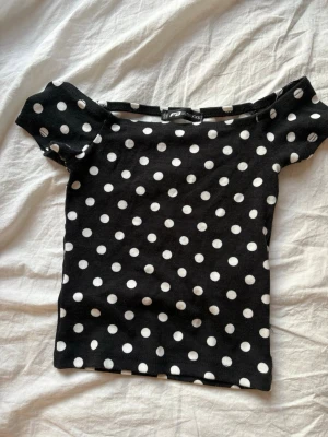 Svart prickig topp  - Trendig svart topp med vita prickar från newyoker. Modellen har korta ärmar och bred, rak urringning som ger en cool retrovibe. Perfekt att styla med jeans eller kjol för en lekfull look.