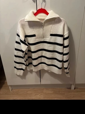 Vit stickad tröja med svarta ränder - Säljer en vit stickad tröja med svarta horisontella ränder. Tröjan har en hög krage med dragkedja och ribbade muddar vid ärmslut och nederkant. Perfekt för dig som gillar klassisk och avslappnad stil.