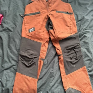 RVRC Nordwand Pro byxor brun/svart - Säljer ett par RVRC Nordwand Pro byxor i brun och svart. Byxorna har förstärkta partier på knän och bak, flera dragkedjeförsedda fickor och elastisk midja. Perfekta för utomhusaktiviteter och äventyr. Materialet är slitstarkt och designen är funktionell.