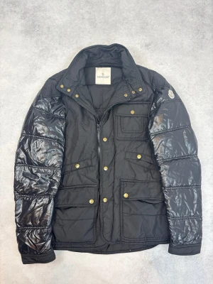 Svart dunjacka från Moncler - Snygg svart dunjacka från Moncler med glansiga ärmar, guldfärgade knappar och flera praktiska fickor framtill. Jackan har hög krage och Moncler-logga på ena ärmen. Perfekt för kalla vinterdagar och riktigt trendig look.