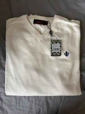 Vit stickad tröja från Morris - Helt ny vit stickad tröja från Morris med V-ringning och broderad blå lilja på bröstet. Tröjan har en klassisk och stilren design, perfekt för lager-på-lager. Materialet är mjukt och bekvämt, vilket gör den enkel att bära hela dagen. Herrstolek-XS. Prislapp kvar! Kan gå ner i pris vid enkel och snabb affär!