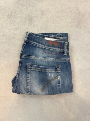 Blå slitna jeans från Dondup - Säljer ett par blå jeans från Dondup med slitningar och tvättade detaljer för en cool, avslappnad look. Jeansen har klassisk femficksdesign, raka ben och kontrastsömmar. Perfekta för dig som gillar streetwear och en trendig stil.