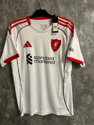 Liverpool FC vit fotbollströja Adidas - Storlek L - Snygg vit matchtröja från Liverpool FC med röda detaljer och Adidas-logga. Tröjan har klubbmärke på bröstet, sponsortryck och röda ränder på axlarna. Tillverkad i lätt och ventilerande material, perfekt för träning eller match.