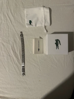 Lacoste silverfärgat klockarmband - Snyggt klockarmband från Lacoste i silverfärgat rostfritt stål. Armbandet har en klassisk länkdesign och pryds av Lacostes ikoniska krokodillogga på spännet. Levereras med originalask och tygpåse med logga.