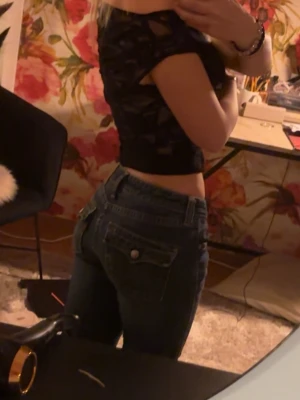 Mörkblå raka jeans med fickdetaljer - Säljer ett par mörkblå jeans med raka ben och klassiska fickor bak med knappdetalj. Jeansen har normal midja och stängs med dragkedja och knapp framtill. Perfekta till en trendig vardagslook.