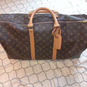 Louis Vuitton monogram resväska - Säljer en klassisk Louis Vuitton resväska i brunt monogrammönster med ljusbruna skinnhandtag och detaljer. Väskan har guldiga metalldetaljer, dragkedja och ikoniska LV-loggan över hela ytan. Perfekt för dig som vill resa med stil och visa upp en tidlös design.