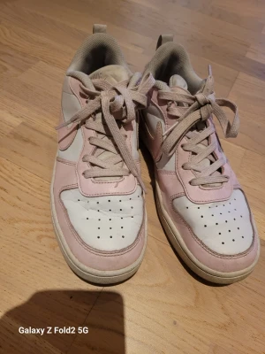 Nike rosa sneakers - Snygga sneakers från Nike i vitt och ljusrosa med klassisk swoosh på sidan. Skorna har lågt skaft, perforerad tåbox och matchande ljusrosa snören. Ovandelen är i läder och textil, vilket ger en fräsch och sportig look.