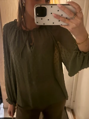 Olivgrön blus med knytning i halsen - Snygg olivgrön blus med diskret prickstruktur och knytning vid halsringningen. Blusen har långa ärmar med resår och volangdetalj vid ärmslut. Tunt, lätt och flowy material som ger en avslappnad look.
