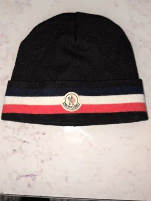 Svart Moncler mössa med ränder - Svart Moncler mössa med klassiska ränder i blått, vitt och rött längs kanten samt Moncler-logga framtill. Snygg och stilren design som passar perfekt till vintern. Materialet är mjukt och bekvämt, perfekt för kalla dagar.