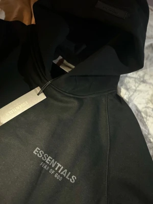 Svart Essentials Fear of God hoodie - Snygg svart hoodie från Essentials Fear of God med tryckt logga på bröstet och ärmen. Klassisk känguruficka framtill och stor huva. Tillverkad i mjukt bomullsmaterial som känns riktigt skönt mot huden. Perfekt för dig som gillar streetwear och vill ha en clean look.