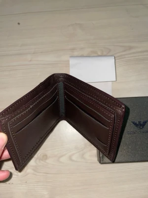 Brun läderplånbok från Armani - Snygg brun plånbok i äkta läder från Giorgio Armani. Plånboken har flera kortfack och ett större fack för sedlar. Klassisk vikbar design med diskret logga på framsidan. Perfekt för dig  som vill ha stilren och praktisk accessoar.