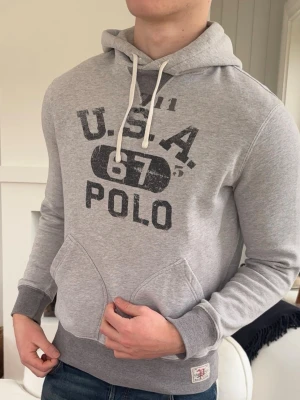 Grå hoodie från Polo Ralph Lauren  - Snygg grå hoodie med tryck 'U.S.A. 67 POLO' på bröstet och vita dragsnören i huvan. Mjuk bomullsblandning, känguruficka framtill och ribbade muddar vid ärmslut och midja. Perfekt för en avslappnad streetstil.