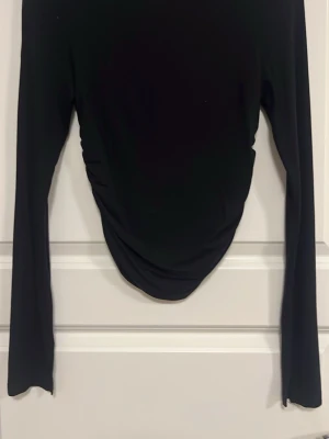 Svart långärmad topp med rynk - Trendig svart långärmad topp med rynkade detaljer framtill. Figurnära passform och rund halsringning. Perfekt att styla med jeans eller kjol för en snygg look.