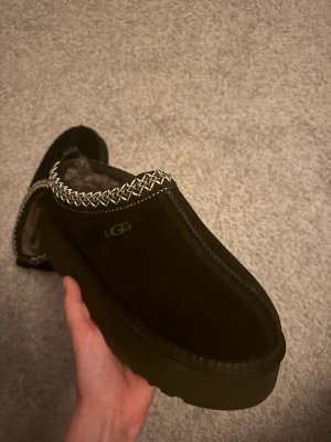 Svarta UGG tofflor med fårskinn - Säljer ett par svarta UGG tofflor i mocka med fluffigt fårskinnsfoder och grov sula. Dekorativt mönster i vitt runt öppningen och diskret UGG-logga på sidan. Perfekta för att hålla fötterna varma och sköna hemma.