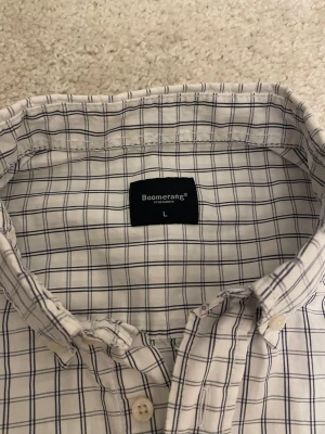 Vit rutig skjorta från Boomerang - Vit skjorta från Boomerang med blått rutmönster. Klassisk krage, knäppning framtill och långa ärmar med manschett. Skjortan är tillverkad i bomull och har en tidlös, stilren look.