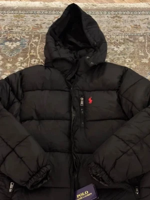 Svart pufferjacka från Polo Ralph Lauren - Snygg svart pufferjacka från Polo Ralph Lauren med huva och dragkedja framtill. Jackan har tre fickor med dragkedja och den klassiska röda loggan broderad på bröstet. Perfekt för kalla dagar