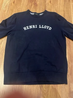 Marinblå sweatshirt från Henri Lloyd - Snygg marinblå stickad tröja från Henri Lloyd med vit broderad logga på bröstet. Tröjan har rund halsringning och ribbade muddar vid ärmslut och nederkant. Perfekt för en avslappnad och stilren look. Står strl xl men sitter som M-L. Skriv för mer bilder