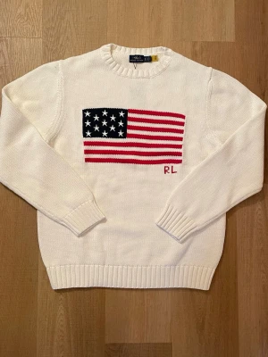 Vit stickad tröja med USA-flagga RL - Vit stickad tröja från Ralph Lauren med en stor USA-flagga i rött, vitt och blått på bröstet samt RL-broderi. Tröjan har rund hals, långa ärmar och ribbstickade muddar vid ärmslut och nederkant. Perfekt för dig som gillar klassisk och cool stil.