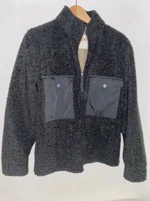 Svart teddyjacka med fickor - Mysig svart teddyjacka med dragkedja framtill och två stora bröstfickor i kontrasterande tyg. Jackan har hög krage och en fluffig, mjuk struktur som ger en avslappnad look. Perfekt för att hålla värmen under kyliga dagar.