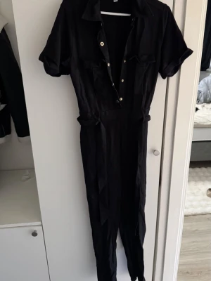 Svart jumpsuit med knappar och skärp - Stilren svart jumpsuit med korta ärmar, krage och knappar framtill. Har två bröstfickor och ett matchande tygskärp i midjan för en snygg siluett. Materialet ser ut att vara mjukt och lätt, perfekt för en trendig look.