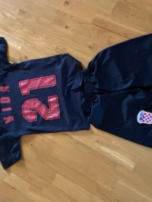 Kroatien fotbollströja och shorts - Säljer ett matchställ med Kroatiens landslagsdesign, svart och mörkblå rutmönstrad tröja med Kroatiens emblem på bröstet och nummer 21 samt namnet Vida på ryggen. Tillhörande svarta shorts med emblem. Perfekt för fotbollsträning eller match. Storlek 164