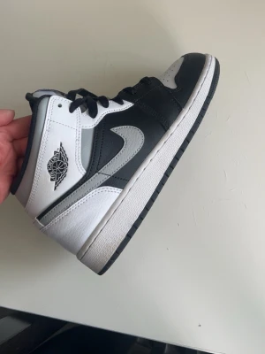 Nike Air Jordan 1 Mid svart/vit/grå - Snygga Nike Air Jordan 1 Mid sneakers i svart, vitt och grått läder. Klassisk siluett med svart swoosh, vita paneler och grå detaljer. Ikonisk Air Jordan-logga på sidan och svart yttersula. Perfekta för dig som gillar streetwear och sportig stil.