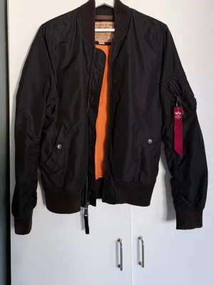 Svart bomberjacka från Alpha Industries - Klassisk svart bomberjacka från Alpha Industries med orange foder, ribbade muddar och dragkedja framtill. Jackan har fickor med tryckknappar och en ikonisk röd tag på ärmen. Perfekt för en cool streetstil.