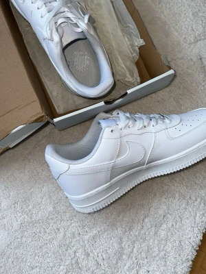 Nike Air Force 1 vita sneakers - Klassiska Nike Air Force 1 sneakers i helvitt läder med vit sula och vita skosnören. Ikonisk design med perforerad tå och tydlig Swoosh-logga på sidan. Perfekta för en clean och stilren look.