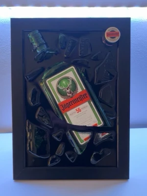 Tavla med krossad Jägermeister-flaska - Unik tavla med en krossad Jägermeister-flaska och kapsyl inramad i svart ram. Flaskan och glasskärvorna är arrangerade konstnärligt mot en svart bakgrund. Perfekt som dekoration för hemmabaren eller som cool inredningsdetalj. Tavlan är egenskapad av Epoxy Creations och gjord av återbruk!🤩