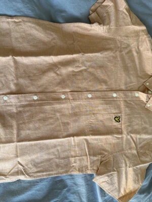 Beige kortärmad skjorta Lyle & Scott - Snygg beige skjorta från Lyle & Scott med korta ärmar och klassisk krage. Skjortan har vita knappar framtill med text loggan på och en bröstficka med den ikoniska gula örnloggan. Perfekt för en avslappnad och stilren look.