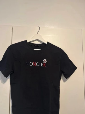 Svart t-shirt med logga - Snygg svart t-shirt från med logga och text i vitt, blått och rött på brösten. som känns skön mot huden. Perfekt för en clean och stilren look. Säljer den eftersom den inte kommer till användning 