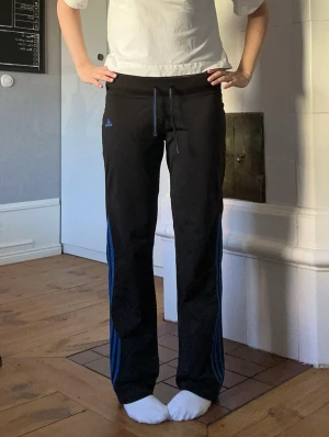 Svarta Adidas mjukisbyxor med blå ränder - Säljer ett par svarta mjukisbyxor från Adidas gamla modell med blå ränder längs benen och dragsko i midjan. Byxorna har raka ben och Adidas-logga på vänster sida. Tillverkade i mjukt och bekvämt material, perfekta för träning eller chill.
