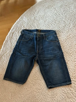 Blå jeansshorts från Lee - Snygga blå jeansshorts från Lee med klassisk femficksdesign och normal passform. Shortsen har medellångt ben och är tillverkade i slitstark bomull. Perfekta för sommardagar och enkel att matcha med t-shirt eller hoodie. Bara att höra av dig för fler frågor. Inga skador på jeansshortsen. Storlek 28