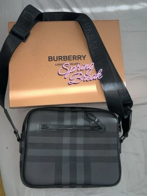 Burberry bag - Snygg svart axelväska från Burberry med klassiskt rutmönster i grå toner. Väskan har justerbar axelrem med Burberry-logga, en dragkedjeficka fram och en öppen ficka bak. Tillverkad i tåligt material med stilren design.