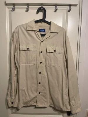 Beige overshirt från Jack & Jones - Stilren beige overshirt från Jack & Jones i mjuk bomull. Skjortan har två bröstfickor med knappar, klassisk krage och långa ärmar med knapp vid manschetten. Perfekt att bära öppen eller stängd för en avslappnad look.