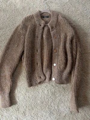 Glittrig guld/beige kofta med knappar - Mysig guld/beige kofta från Gina Tricot med glittriga inslag med knappar framtill. Koftan har en fluffig, stickad struktur och rund halsringning. Perfekt för att addera lite extra stil till din outfit.