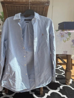 Ljusblå skjorta från Polo Ralph Lauren - Klassisk ljusblå skjorta från Polo Ralph Lauren med broderad logga på bröstet. Skjortan har knappar framtill, lång ärm och en tidlös krage. Perfekt för en clean och stilren look.