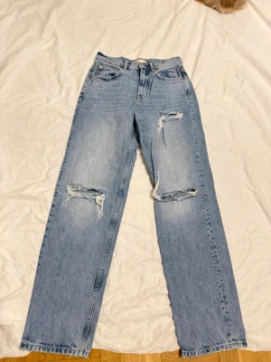 Blå ripped jeans från Gina Tricot - Säljer ett par ljusblå jeans från Gina Tricot med raka ben och slitna detaljer vid knäna. Jeansen har klassisk femficksmodell och hög midja. Perfekta för en avslappnad och trendig look.