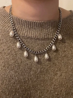 Silverfärgat halsband med pärlor - Snyggt halsband med kraftig silverfärgad kedja och hängande droppformade vita pärlor. Perfekt för att ge en cool och elegant touch till din outfit. Materialet är rostfritt och pärlorna har en glansig finish.