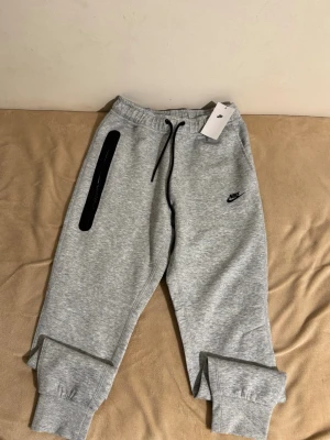 Grå Nike mjukisbyxor med logga - Snygga grå mjukisbyxor från Nike med svart logga på vänster ben. Byxorna har svarta snören i midjan, ribbade muddar vid benslut och en svart detalj längs ena fickan. Perfekt för chill eller träning.