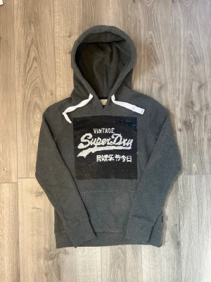 Grå hoodie från Superdry med paljetter - Snygg grå hoodie från Superdry med stor paljettpatch på bröstet där det står 'Vintage Superdry' och japanska tecken🤩 Storlek Xs med inga fel. Hör av er vid funderingar samt snabbaffär😇