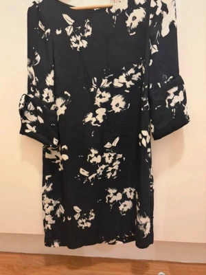 Svartvit blommig klänning - Svart klänning med vita blommor och trekvartsärm. Rak passform och rund halsringning. Ärmarna har en lätt volangdetalj. Perfekt för dig som gillar en stilren men ändå lekfull look.