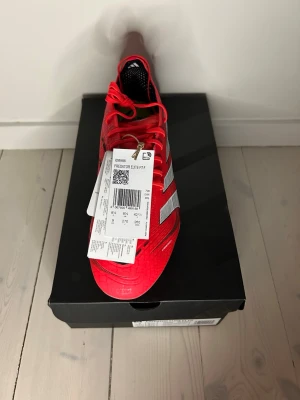 Adidas Predator Elite FT FG - Säljer ett par Adidas Predator Elite FT FG fotbollsskor i rött med vita detaljer och klassiska tre ränder på sidan. Skorna har snörning och FG-dobbar för spel på naturgräs. Snygg, modern design och lättviktigt syntetmaterial för bästa bollkontroll.