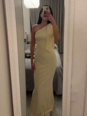Ljusgul one shoulder långklänning - Ljusgul långklänning med one shoulder-design och stor rosettdetalj vid axeln. Klänningen har en figurnära passform. Endast använd en gång och i mycket bra skick. Obs: Dragkedjan kan vara lite trög/har en mindre defekt, men den går att dra upp och fungerar som den ska när man väl får till den. Originalpris: 1 350 kr🌸